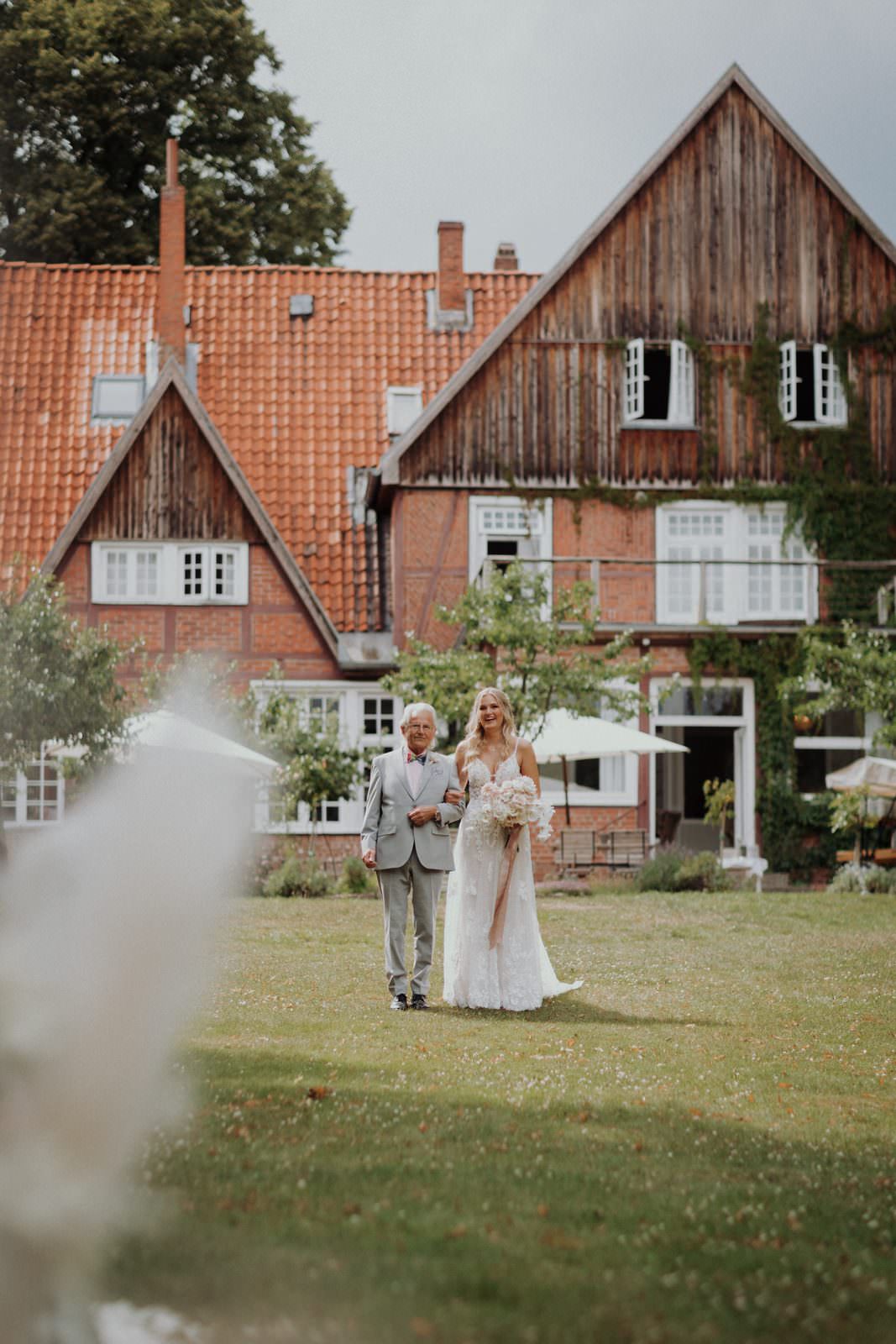 Cyana und ihr Großvater unterwegs zum freien Altar, das Hotel im Hintergrund, Pampasgras unscharf im Vordergrund.